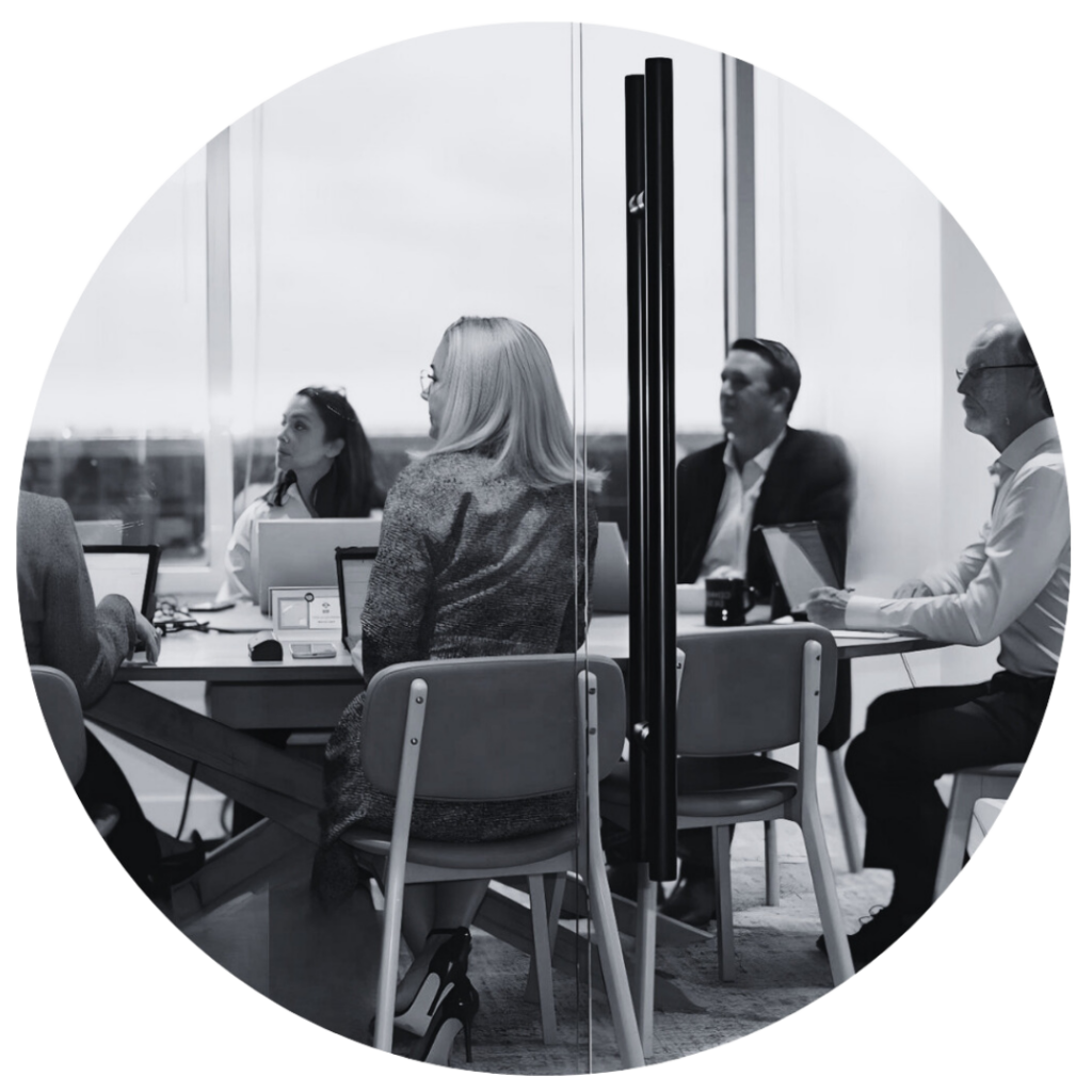 A group of five people on laptops at a boardroom office table listening to a presenter.